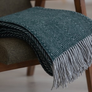May include: A green and white patterned throw blanket with a fringe border. The blanket is folded and draped over a wooden chair.