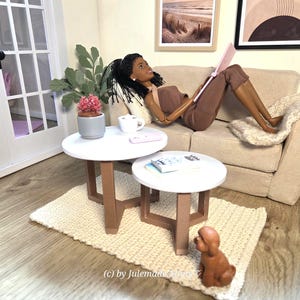 Könnte beinhalten: Eine Miniaturpuppe mit dunkler Haut und geflochtenem Haar in braunem Oberteil und Hose, die auf einem beigen Sofa liegt und einen rosa Laptop hält. Zwei weiße runde Tische mit braunen Beinen, eine kleine Topfpflanze, eine Tasse und ein Hund.