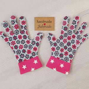 Peut inclure: Une paire de gants de jardinage gris et rose avec un motif floral blanc. Les gants ont des poignets roses avec des étoiles blanches. Les gants sont faits à la main et l'étiquette indique "handmade Julemade".