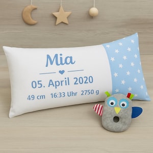 Könnte beinhalten: Ein weißes Kissen mit blauem Sternenmuster auf einer Seite. Das Kissen ist personalisiert mit dem Namen "Mia", dem Datum "05. April 2020", der Uhrzeit "16:33 Uhr" und dem Gewicht "2750 g". Das Kissen ist 49 cm lang. Ein grauer Eulenrassel mit bunten Akzenten befindet sich im Vordergrund.