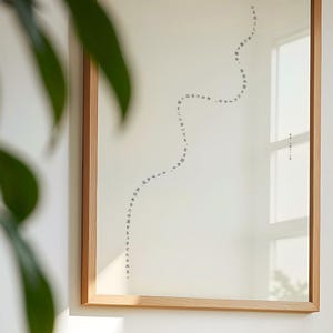 Puede incluir: Una impresión enmarcada con un marco de madera clara. La impresión presenta texto japonés en una línea curva sobre un fondo blanco. El texto es negro. El marco es rectangular y cuelga de una pared blanca.