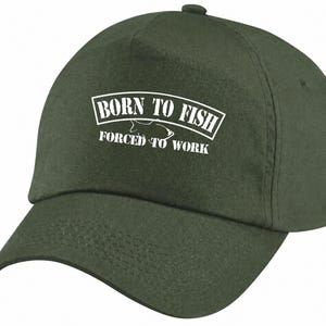 May include: An olive green baseball cap featuring white embroidered text that reads BORN TO FISH FORCED TO WORK with a small fish graphic positioned between the two lines of text. The cap has a curved brim and a structured crown design.