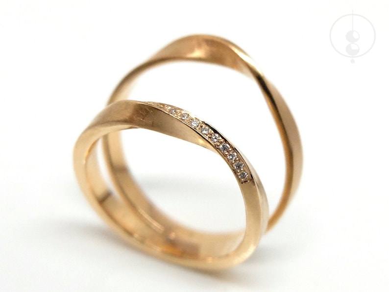 Nahaufnahme von zwei Möbius-Ringen aus Roségold, die seitlich versetzt nebeneinander auf weißem Untergrund stehen. Beide Ringe haben eine Drehung. Im vorderen, kleineren Ring sind im oberen Drittel der Drehung viele kleine Diamanten gefasst.