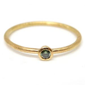 May include: A thin gold ring featuring a single, small, round, bezel-set green gemstone. The band has a smooth, polished finish, and the setting securely holds the dark green stone in the center, creating a simple and refined piece of jewelry.
