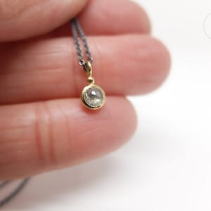 Unterer Teil einer geschwärzten Silberkette mit einem runden Anhänger aus Gold mit einer grauen Diamantrose. Zwischen Anhängeröse und Fassung sitzt ein kleines Goldkügelchen. Der Anhänger liegt auf dem kleinen Finger einer Hand.