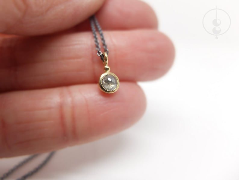 Unterer Teil einer geschwärzten Silberkette mit einem runden Anhänger aus Gold mit einer grauen Diamantrose. Zwischen Anhängeröse und Fassung sitzt ein kleines Goldkügelchen. Der Anhänger liegt auf dem kleinen Finger einer Hand.