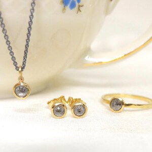 Nahaufnahme von einem Schmuckset aus Gold mit  grauen Diamanten im Rosenschliff. Das Set besteht aus einer geschwärzten Silberkette mit Anhänger, einem Paar Ohrstecker und einem Ring.  Die Kette hängt auf dem Rand einer alten Goldrandtasse mit blauen Blümchen. Die Ohrringe und der Ring liegen rechts auf dem Boden der Tasse.