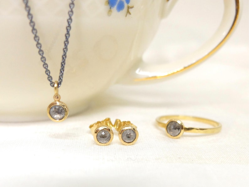 Nahaufnahme von einem Schmuckset aus Gold mit  grauen Diamanten im Rosenschliff. Das Set besteht aus einer geschwärzten Silberkette mit Anhänger, einem Paar Ohrstecker und einem Ring.  Die Kette hängt auf dem Rand einer alten Goldrandtasse mit blauen Blümchen. Die Ohrringe und der Ring liegen rechts auf dem Boden der Tasse.