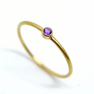 Könnte beinhalten: Ein dünner Goldring mit einem einzelnen, kleinen, runden, violetten Edelstein in einer Zargenfassung. Die Ringschiene hat eine glatte, matte Oberfläche und ein schlichtes, kreisförmiges Design. Der violette Stein ist oben auf dem Ring zentriert.