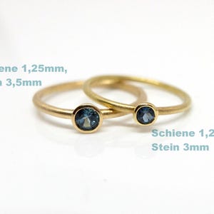 以下が含まれることがあります： 青い宝石が付いた2つのゴールドリング。大きい方のリングは3.5mmの石と1.25mmのバンドが付いています。小さい方のリングは3mmの石と1.25mmのバンドが付いています。リングは白い背景にあります。