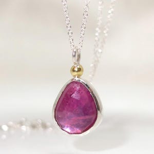 Könnte beinhalten: Eine silberne Halskette mit einem Anhänger mit einem großen, facettierten, rosa Edelstein. Der Anhänger ist tropfenförmig und in eine silberne Lünette gefasst. Eine kleine goldene Perle verbindet den Anhänger mit der Silberkette.