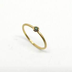 May include: A thin gold ring featuring a single, small, round, bezel-set green gemstone. The band has a smooth, matte finish, and the setting securely holds the circular stone in place, creating a simple and refined piece of jewelry.
