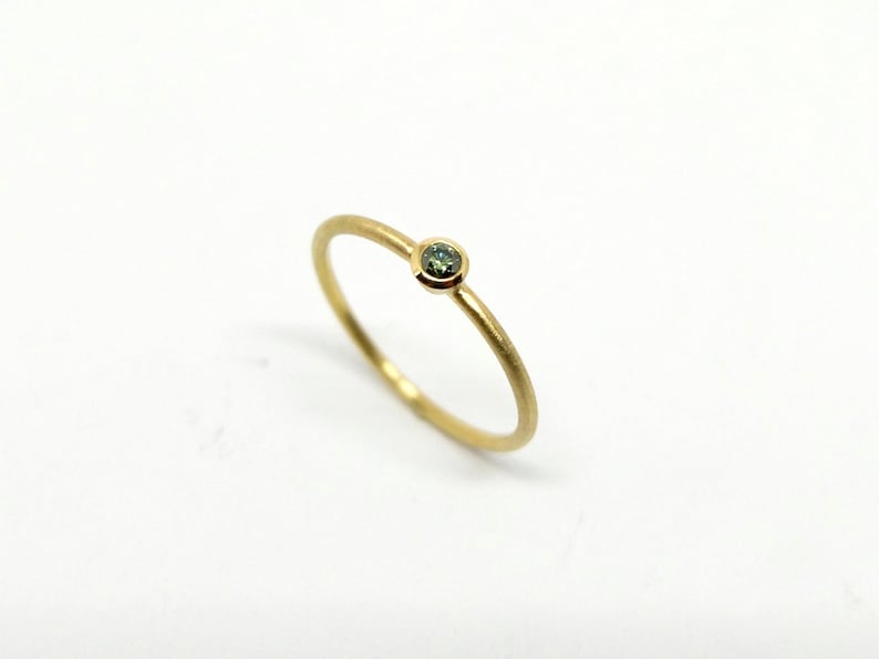 May include: A thin gold ring featuring a single, small, round, bezel-set green gemstone. The band has a smooth, matte finish, and the setting securely holds the circular stone in place, creating a simple and refined piece of jewelry.