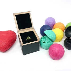 May include: A square black wooden ring box with a light-colored rim containing a ring with a round green stone. Beside it are a red heart-shaped box and several small, colorful, textured spherical containers, one of which holds a thin silver ring.
