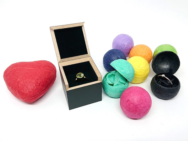 May include: A square black wooden ring box with a light-colored rim containing a ring with a round green stone. Beside it are a red heart-shaped box and several small, colorful, textured spherical containers, one of which holds a thin silver ring.