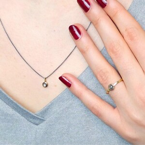 Puede incluir: Un delicado collar de cadena de plata con un peque&ntilde;o colgante redondo con una piedra preciosa transparente. Un anillo de oro con una piedra preciosa similar se lleva en el dedo &iacute;ndice de una mano con esmalte de u&ntilde;as rojo.