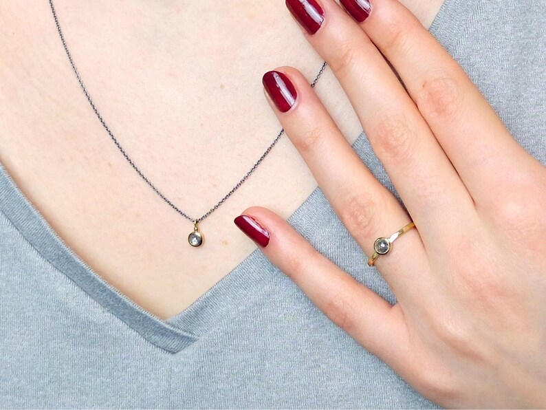 Puede incluir: Un delicado collar de cadena de plata con un peque&ntilde;o colgante redondo con una piedra preciosa transparente. Un anillo de oro con una piedra preciosa similar se lleva en el dedo &iacute;ndice de una mano con esmalte de u&ntilde;as rojo.