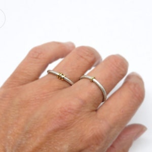 Aufnahme von linker Frauenhand, an der am Ringfinger und am Mittelfinger je ein Silberring mit runder Ringschiene und matter Oberfläche sitzen. An dem einen Ring sitzen 3 facettierte Goldösen, am anderen Ring nur eine Öse.