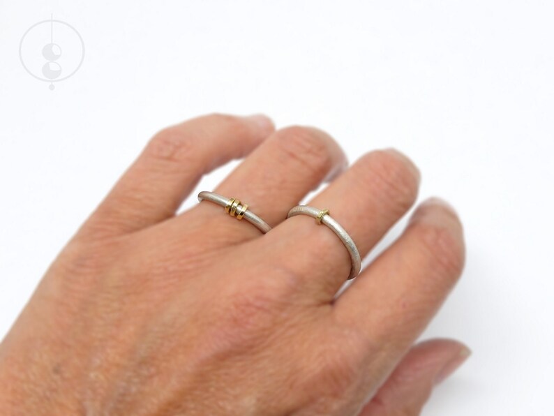Aufnahme von linker Frauenhand, an der am Ringfinger und am Mittelfinger je ein Silberring mit runder Ringschiene und matter Oberfläche sitzen. An dem einen Ring sitzen 3 facettierte Goldösen, am anderen Ring nur eine Öse.