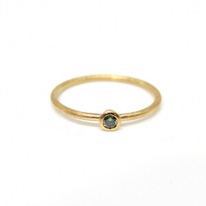 May include: A thin gold ring featuring a single, small, round-cut dark green gemstone set in a bezel setting. The band has a matte, textured finish, and the gemstone is positioned at the center of the ring, creating a simple and delicate appearance.