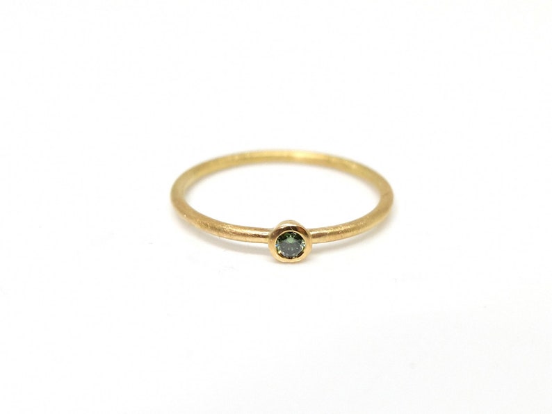 May include: A thin gold ring featuring a single, small, round-cut dark green gemstone set in a bezel setting. The band has a matte, textured finish, and the gemstone is positioned at the center of the ring, creating a simple and delicate appearance.