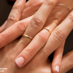 Nahaufnahme von zwei ineinander verschränkten Händen, die jeweils einen Möbius-Ringen aus Roségold tragen. Beide Ringe haben eine Drehung, deren Kanten poliert sind.