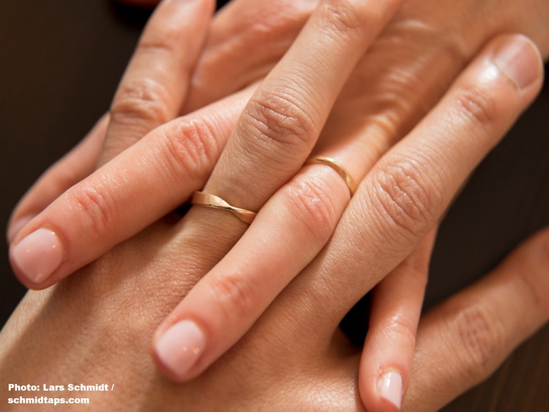 Nahaufnahme von zwei ineinander verschränkten Händen, die jeweils einen Möbius-Ringen aus Roségold tragen. Beide Ringe haben eine Drehung, deren Kanten poliert sind.