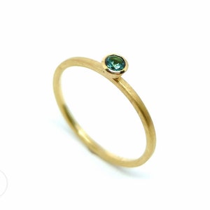K&ouml;nnte beinhalten: Ein schlichter Goldring mit einem kleinen, runden, blaugr&uuml;nen Edelstein in der Mitte.
