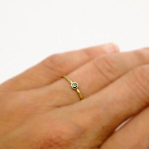 May include: A thin gold band ring featuring a single, small, round, bezel-set green gemstone. The ring has a polished metallic finish and a simple, slender design that sits on the finger, showcasing the contrast between the gold and stone.