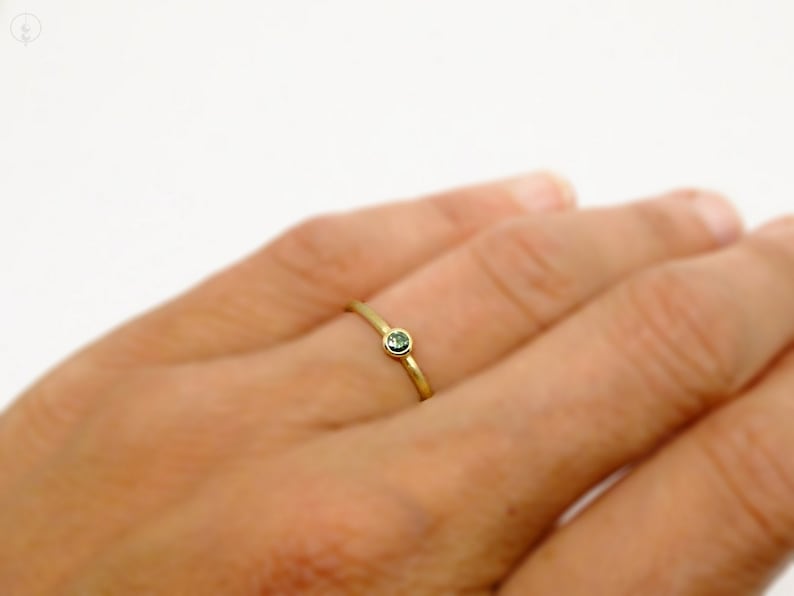 May include: A thin gold band ring featuring a single, small, round, bezel-set green gemstone. The ring has a polished metallic finish and a simple, slender design that sits on the finger, showcasing the contrast between the gold and stone.