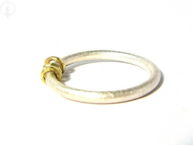 Silberring auf weißem Untergrund mit matter Oberfläche. Die Ringschiene ist aus 2mm dickem Runddraht angefertigt und der Ring mit drei verschieden facettierte Goldösen verziert, die seitlich zum Betrachter ausgerichtet nebeneinander liegen.