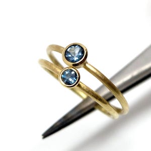 Nahaufnahme von zwei goldenen Ringen mit hellblauen Steinen, die von einer Pinzette vor weißem Untergrund gehalten werden. Die Ringe haben eine matte Oberfläche und eine runde Fassung, die etwas über die Ringschiene hinausragt. Die Steine der beiden Ringe sind unterschiedlich groß - der untere ist etwas kleiner.