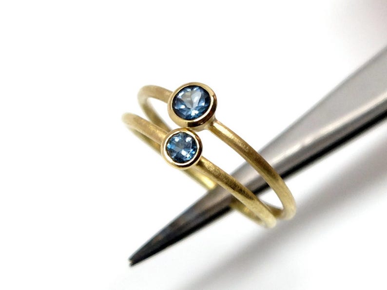Nahaufnahme von zwei goldenen Ringen mit hellblauen Steinen, die von einer Pinzette vor weißem Untergrund gehalten werden. Die Ringe haben eine matte Oberfläche und eine runde Fassung, die etwas über die Ringschiene hinausragt. Die Steine der beiden Ringe sind unterschiedlich groß - der untere ist etwas kleiner.