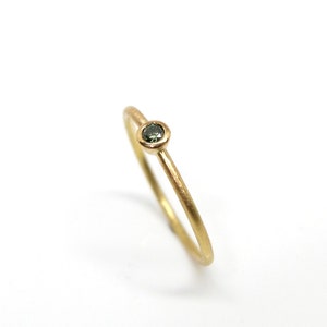 May include: A thin gold ring with a matte finish featuring a single, small, round, dark green gemstone set in a simple bezel setting. The band is slender and circular, showcasing a clean, understated design with a polished interior surface.