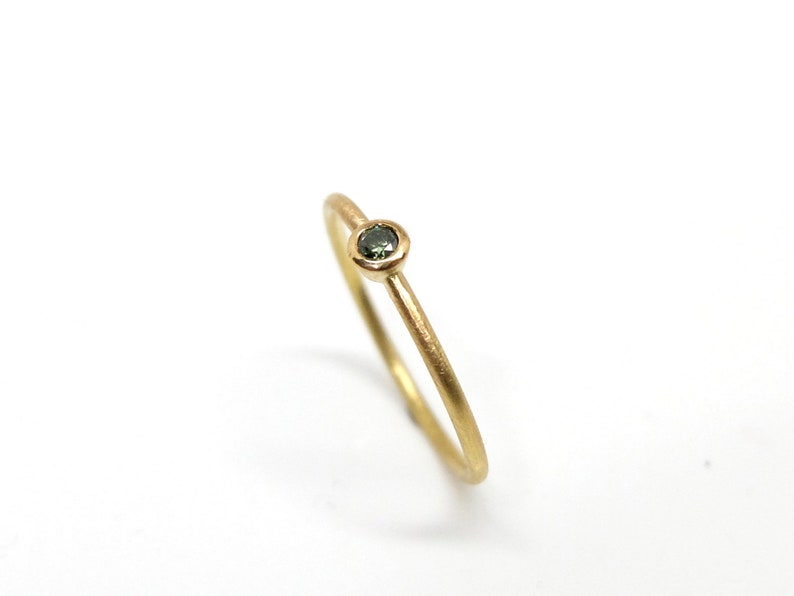 May include: A thin gold ring with a matte finish featuring a single, small, round, dark green gemstone set in a simple bezel setting. The band is slender and circular, showcasing a clean, understated design with a polished interior surface.