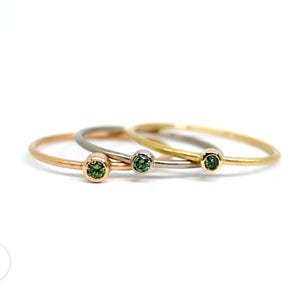 Drei schmale Ringe mit grünen Diamanten in unterschiedlichen Goldlegierungen liegen seitlich versetzt übereinander auf weißem Untergrund. Von links nach rechts erst Rotgold, dann Weißgold und ganz rechts Gelbgold.