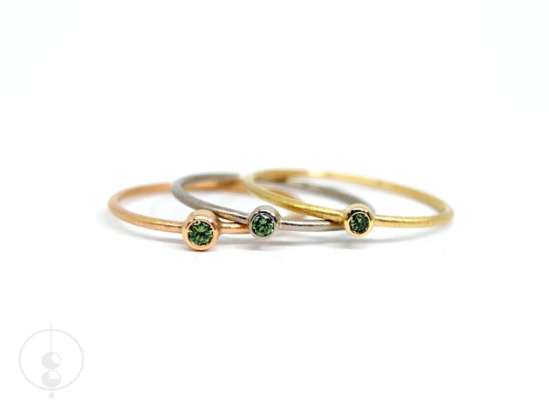 Drei schmale Ringe mit grünen Diamanten in unterschiedlichen Goldlegierungen liegen seitlich versetzt übereinander auf weißem Untergrund. Von links nach rechts erst Rotgold, dann Weißgold und ganz rechts Gelbgold.