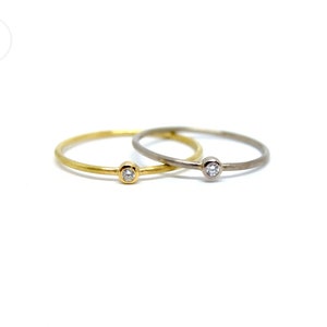 Zwei dünne Ringe, einer aus Weißgold, einer aus Gelbgold, beide mit kleinem Diamanten in runder Zargenfassung. Die Ringe haben eine matte Oberfläche und liegen auf weißem Untergrund mit der Fassung nach vorne zum Betrachter.
