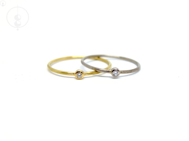 Zwei dünne Ringe, einer aus Weißgold, einer aus Gelbgold, beide mit kleinem Diamanten in runder Zargenfassung. Die Ringe haben eine matte Oberfläche und liegen auf weißem Untergrund mit der Fassung nach vorne zum Betrachter.