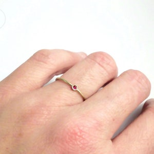 Dünner Goldring mit matter Oberfläche, der auf Ringfinger einer linken Hand vor weißem Hintergrund. Im Ring gefasst ist ein kleiner, pinkroter Rubin.