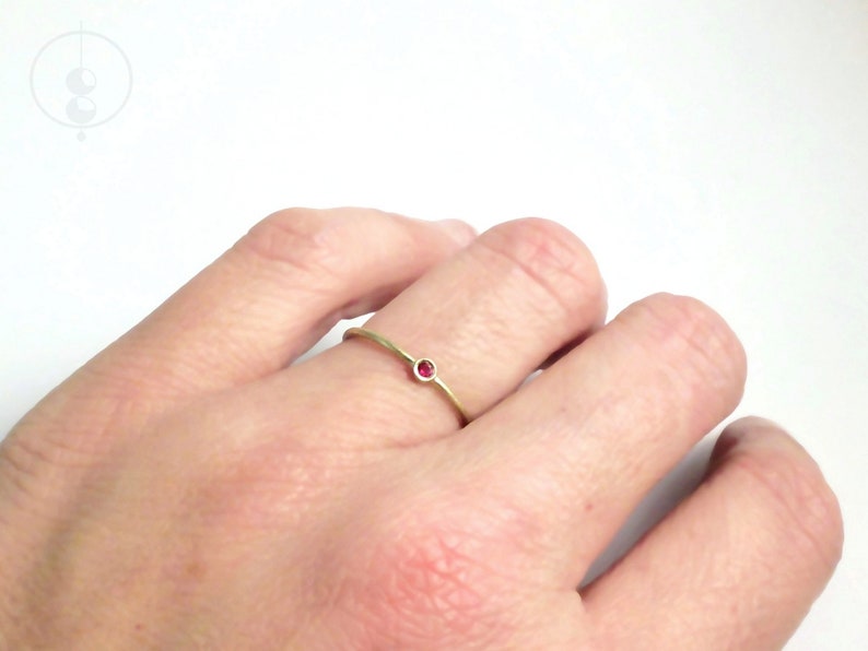 Dünner Goldring mit matter Oberfläche, der auf Ringfinger einer linken Hand vor weißem Hintergrund. Im Ring gefasst ist ein kleiner, pinkroter Rubin.