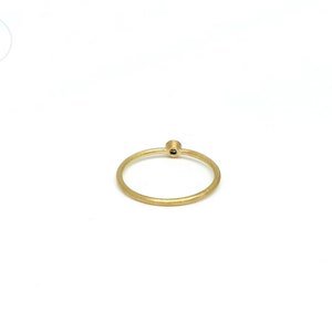 May include: A thin, circular gold ring with a matte, textured finish. The band features a single, small, dark green gemstone set in a raised, cylindrical gold setting positioned at the top of the ring, creating a simple and delicate appearance.