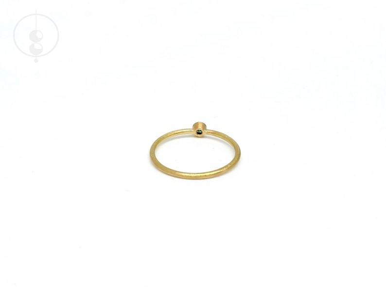 May include: A thin, circular gold ring with a matte, textured finish. The band features a single, small, dark green gemstone set in a raised, cylindrical gold setting positioned at the top of the ring, creating a simple and delicate appearance.