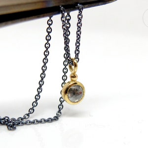 Unterer Teil einer geschwärzten Silberkette mit einem runden Anhänger aus Gold mit einer grauen Diamantrose. Zwischen Anhängeröse und Fassung sitzt ein kleines Goldkügelchen. Die Kette wird von Pinzette vor weißem Hintergrund gehalten.