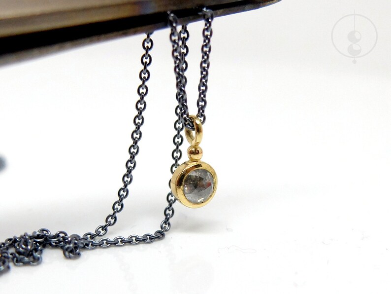 Unterer Teil einer geschwärzten Silberkette mit einem runden Anhänger aus Gold mit einer grauen Diamantrose. Zwischen Anhängeröse und Fassung sitzt ein kleines Goldkügelchen. Die Kette wird von Pinzette vor weißem Hintergrund gehalten.