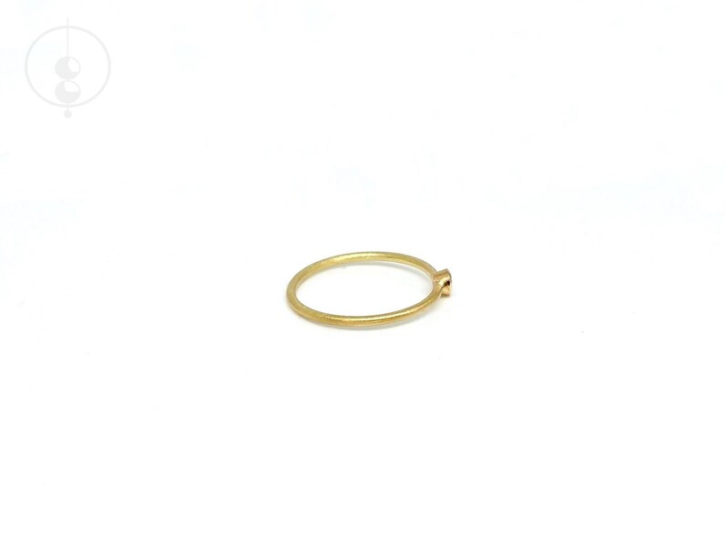 May include: A thin, circular gold ring with a matte, brushed texture. The band is uniform in thickness and features a single, small, round, bezel-set stone at the top. The metal has a warm, yellow hue and a subtle, non-reflective finish.