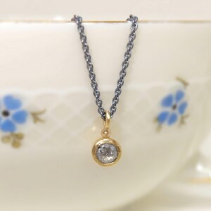 Nahaufnahme von dem unteren Teil einer geschwärzten Silberkette mit einem runden Anhänger aus Gold, in dem einer kleiner, grauer Diamant im Rosenschliff gefasst ist. Zwischen Anhängeröse und Fassung sitzt ein kleines Goldkügelchen. Die Kette hängt auf dem Rand einer alten Goldrandtasse mit blauen Blümchen.