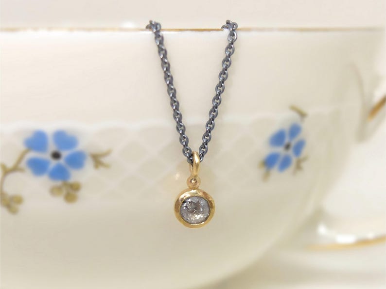 Nahaufnahme von dem unteren Teil einer geschwärzten Silberkette mit einem runden Anhänger aus Gold, in dem einer kleiner, grauer Diamant im Rosenschliff gefasst ist. Zwischen Anhängeröse und Fassung sitzt ein kleines Goldkügelchen. Die Kette hängt auf dem Rand einer alten Goldrandtasse mit blauen Blümchen.