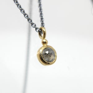 Nahaufnahme von dem unteren Teil einer geschwärzten Silberkette mit einem runden Anhänger aus Gold, in dem einer kleiner, grauer Diamant im Rosenschliff gefasst ist. Zwischen Anhängeröse und Fassung sitzt ein kleines Goldkügelchen.