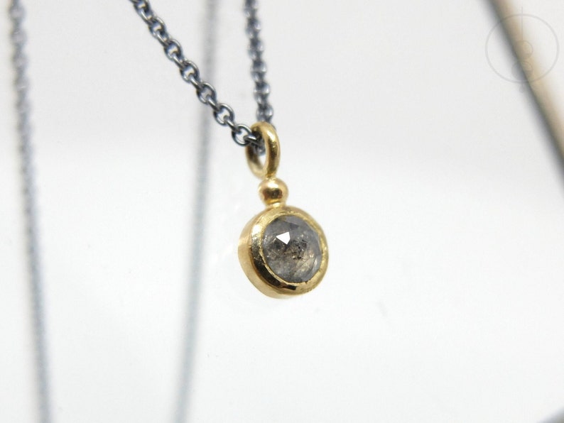 Nahaufnahme von dem unteren Teil einer geschwärzten Silberkette mit einem runden Anhänger aus Gold, in dem einer kleiner, grauer Diamant im Rosenschliff gefasst ist. Zwischen Anhängeröse und Fassung sitzt ein kleines Goldkügelchen.